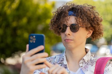 Mutlu, gülümseyen esmer adam internet sosyal medya uygulamalarını karıştırarak açık havada sohbet ederek akıllı telefon yazıları yazıyor. Şehir sokağında kıvırcık saçlı bir turist. Yaşam Tarzları.