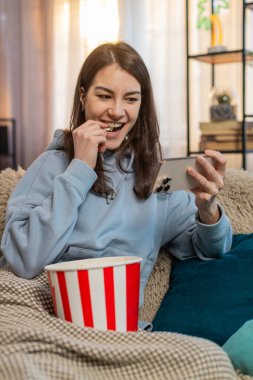 Mutlu Kafkasyalı gevşemiş kadın evinde oturup akıllı telefondan film izleyip patlamış mısır yiyor. Üstü başı battaniyeyle örtülü genç bayan oturma odasında bir dizi izlerken mutlu oluyor..