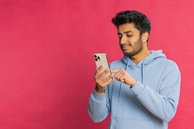 Mutlu Hintli genç adam sosyal medyada geziniyor, akıllı telefona bakarken gülümsüyor, ilginç içeriğin tadını çıkarıyor ve bir mesaj yazıyor. Çevrimiçi bağlanan rahat Arap adam, kırmızı arka planda izole edilmiş.