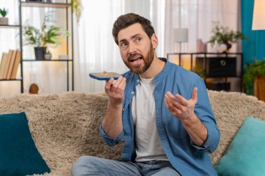 Evdeki genç adam akıllı telefondan bir sesli mesajı dikkatle dinliyor, başını eğiyor ve konsantre oluyor. Kafkasyalı adam koltukta oturmuş, net bir şekilde duymaya çalışıyor ve önemli ses detaylarına odaklanıyor..