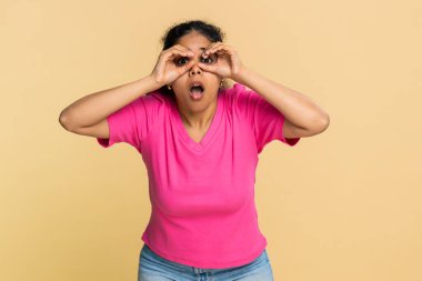 Hintli kadın göz çevresinde şakacı bir şekilde parmak bardakları oluşturur. Bej arka planda izole edilmiş Arap kızı esprili bir şekilde merak, oyuncu bir ruh hali, yaratıcılık, eğlence ve dostça bir etkileşim sergiliyor..