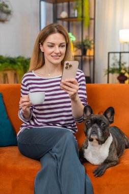 Orta yaşlı bir kadın evde Fransız buldoguyla kahve içiyor. Akıllı telefondan haberleri okuyor. Sakin ve mutlu hissediyor. Kız evcil köpek kanepeye oturmuş gülümsüyor. Kupanın keyfini çıkarıyor. Sabah haberlerine bakıyor. Günlük yaşam tarzını internette tarıyor.