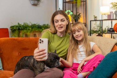 Evde Fransız bulldog 'u olan anne ve kız akıllı telefon şakası gülüşünde selfie filtre maskesi deniyor. Kanepede köpekli kadın ve kız eğlenceli fotoğraf çekiyorlar. Maskelerle oynuyorlar. Sıcak bir anı paylaşıyorlar.
