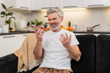 Evde yaşayan orta yaşlı bir adam akıllı telefon dalgalarında ses kaydı yapıyor merhaba hoparlörden ve haberleri paylaşıyor. Kanepede gülümseyen olgun bir adam konuşuyor sohbet yanıtı gönderiyor sonra da dinletiyor ve kısa bir güncelleme ekliyor..