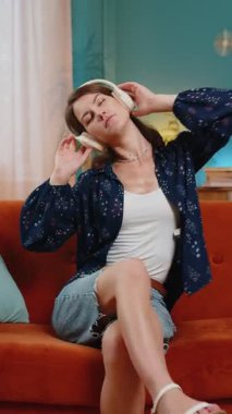 Kanepede uzanmış, kablosuz kulaklıkla müzik dinleyen neşeli genç bir kadın. Evde müzik eşliğinde dans eden sıradan giyinmiş neşeli yetişkin bir kız. Dans müziğinin tadını çıkarıyorum. Yaşam Tarzları.