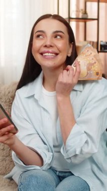 Evde elinde euro parası ve akıllı telefonu olan genç bir kadın parasını nereye harcayacağına karar veriyor. Kanepede oturan beyaz kız dikkatli bir şekilde finans, bütçe ve satın alma seçeneklerini değerlendiriyor.