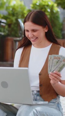 Mutlu zengin kadın dizüstü bilgisayar kazananı zaferini kullanıyor. Nakit para tutuyor. Açık havada gündüz vakti sokakta oturuyor. Genç esmer kız turist yatırım bütçesi planlama başarısı.