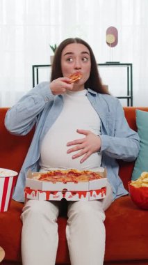 Sağlıksız abur cubur pizza yiyen hamile bir kadın, patlamış mısır gevreği kanepede oturmuş evde televizyon seyrediyor. Hamilelik süresince dairede yağlı yemekler. Açlık, tat tercihlerindeki değişim.