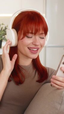 Evde kablosuz kulaklık takan genç bir kadın akıllı telefondan müzik dinliyor ve dinleniyor. Kanepede oturan kızıl saçlı kız gözlerini kapatır yavaşça nefes alır yumuşak bir gülümseme sakin bir ruh hali ve kısa bir dinlenme.