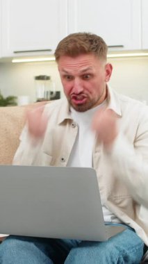 Evde çalışan genç adam dizüstü bilgisayarı kontrol ediyor, proje onayı alıyor ve sevinçle kutluyor. Kanepede oturan beyaz adam elini kaldırıyor zafer jestleri yapıyor gülümsüyor ve iyi haberlerin tadını çıkarıyor ve mutlu yorumlar yapıyor..