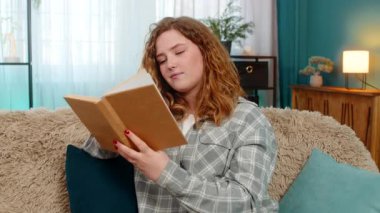 Genç bir kadın evde kitap okuyor ve derin içeriği yansıtırken sayfaları yavaşça çeviriyor. Kanepedeki beyaz saçlı kız düşünceli bir şekilde kitap okuyor ve entellektüel anın tadını çıkarıyor.