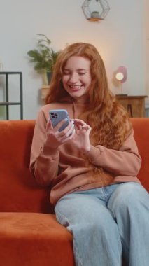 Evdeki oturma odasında gülümseyen cep telefonu kullanan kızıl saçlı mutlu bir genç kadın. Akıllı telefon sosyal medya uygulamalarında internetten film izleyen kız mesajları paylaşıyor.