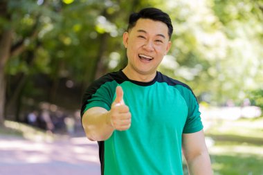 Gibi. Portre atletik formda gülümseyen koşucu eğitimi kameraya bakıp maraton başarısına seviniyor. Parkta egzersiz kardiyolojisi. Japon genç sporcu bulanık ağaçlara karşı baş parmağını kaldırıyor