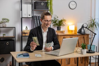 Genç ve mutlu iş adamı para sayıyor ve merkez ofiste dizüstü bilgisayar kullanıyor. Başarılı serbest çalışan, ofise para getiren, temettüleri hesaplayan, masadaki nakit kârları ve mali durumu hesaplayan biri..