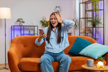 Üzgün, Kafkasyalı bir kadın akıllı telefon daktilosu kullanıyor, aniden piyango sonuçlarını şaşırıyor, kötü haber, servet kaybı, son teslim tarihi, virüs. Evdeki kanepede oturan genç kız.