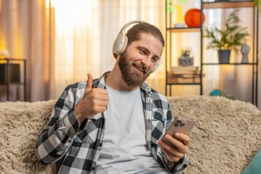 Genç adam kablosuz kulaklıklarla evde titrer müzik dinler, akılda kalıcı bir ritimle başını sallar. Kanepedeki beyaz adam başparmağını kaldırdı, bu da harika bir melodiyi onayladığını gösteriyor.