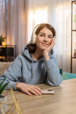 Kanepede oturan, akıllı telefondan sesli telefon dinleyen, kablosuz kulaklık takan beyaz bir kadın. Kız sakin bir odada sakin bir ifadeyle rahatlamış görünüyor. Odaklanmış zihniyle eğleniyor.
