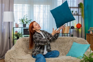 Genç, mutlu bir kadın evinde, neşeli bir gülümsemeyle yastığı fırlatıyor. Kaygısız anın tadını çıkarıyor. Kafkasyalı kızıl kız kanepede eğleniyor, neşeli ruh hali ve duygusal rahatlama gösteriyor..