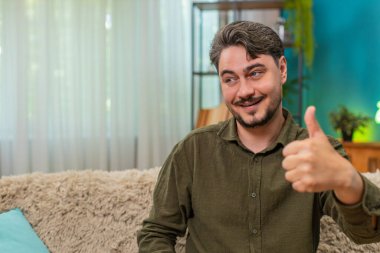 Evde orta yaşlı bir adam olumlu geri bildirim başarısı gibi gülümseyerek başparmağını kaldırır. Kanepedeki genç adam memnuniyetini ifade ediyor. Büyük sonuç jesti neşe gururu özgüvenini yansıtıyor.