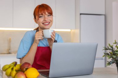 Evde oturan genç bir kadın dizüstü bilgisayarla e-postalarına cevap veriyor ve sıcak kahveyi yudumluyor. Mutfaktaki kızıl saçlı kız gelen kutusuna açık cevaplar yazar notlar düzenler ve iş için sonraki adımları planlar..