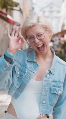 Çekici genç milenyum kızının portresi el işaretini gösteren kameraya bakıyor. Kısa sarı saçlı beyaz kadın her şeyi gösteriyor. Güzel başarı jesti vücut dili. Kameraya gülümseyen kadın.
