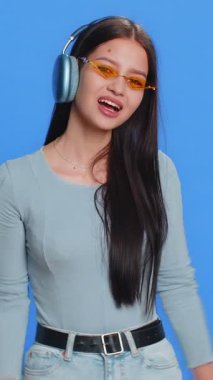 Kulaklık takmış, müzik dinleyen, dans eden, eğlenen, el kol hareketi yapan, partide gevşeyen mutlu bir kadın. Güneş gözlüklü esmer kız stüdyo mavi arka planda izole edilmiş.