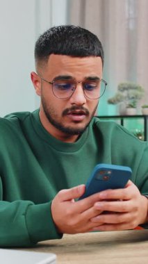 Hüzünlü Arap adam akıllı telefon daktilo kullanıyor, beklenmedik piyango sonuçlarını kaybediyor, kötü haber, servet kaybı, son teslim tarihi, virüs. Masadaki ofisteki genç Hintli adam.