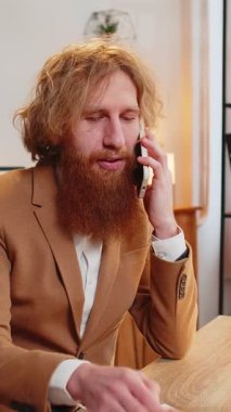 Genç işadamı ofis masasında oturuyor cep telefonuyla müşteriye cevap veriyor, akıllı telefon kullanıyor. Sohbet. Kafkasyalı kızıl saçlı, uzaktan kumandalı bir işte çalışıyor..