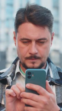 Happy şoke olmuş Kafkasyalı yetişkin turist gündelik giysiler içinde ağzı açık bir şekilde zaferi kutluyor cep telefonlarıyla iyi haberler taşıyor, mesajlar kazanıyor şehir merkezinin dışında başarılar diliyor.