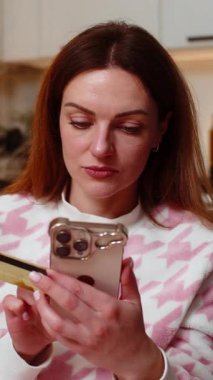 Akşamları evde oturan orta yaşlı bir kadın online siparişleri akıllı telefon ve kredi kartı tebessümleri ile ödüyor. Akşamları kanepede oturan beyaz kız ödeme durumunu doğruladı.