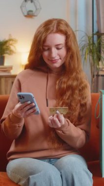 Alışveriş meraklısı kızıl saçlı mutlu bir kadın alışveriş torbalarıyla evde oturuyor online sipariş veriyor, kredi kartı ile ödeme yapıyor. Evdeki koltukta oturan heyecanlı kadın...