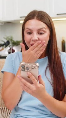 Evdeki genç kadın iş okuyor akıllı telefondan e-posta gönderiyor ağız kapaklarını sevinçle kaplıyor ve rahatlıyor. Kanepedeki beyaz kız kontrat imzaladı ve kabul etti. Uygulamada cevap verdi. Heyecanlı, mutlu, harika haberler.