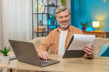 Beyaz, olgun bir adam evde oturmuş önemli belgeleri inceliyor ve dizüstü bilgisayar kullanıyor. Orta yaşlı bir adam başarılı bir işten aldığı memnuniyetle gülümser, verimlilik ve başarıyı gösterir.