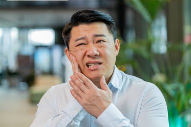 Ofis lobisindeki Çinli işadamı dizüstü bilgisayarla çalışıyor ve diş ağrısının yanağa dokunduğunu hissediyor diş hastalığından dolayı acı veren bir ifade. Pulpitis, sakız hastalığı, diş hastalıkları konsepti.