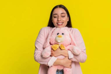 Genç bir kadın, pembe doldurulmuş oyuncak tavşana sarılmış, mutlu, nazik bir gülümsemeyle, sıcaklığını ve şefkatini ifade ediyor. Sarı arka planda izole edilmiş bir kız samimi duygusal tepkiler ve huzurlu neşeli bir ruh hali sergiliyor.