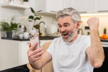 Evde orta yaşlı bir adam akıllı telefondan mutlu haberler okur ve online oyun kutlarken güler başarılı olur neşeli ruh hali kazanır. Kanepede oturan olgun bir adam gülümseyerek yumruk zaferini kaldırıyor..