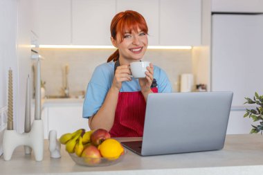 Evde oturan genç bir kadın dizüstü bilgisayarla e-postalarına cevap veriyor ve kahveden bir yudum alıyor. Mutfaktaki kızıl saçlı kız, gelen kutusuna cevap veriyor. Müşteriler için taslak notlar. İşine odaklan..