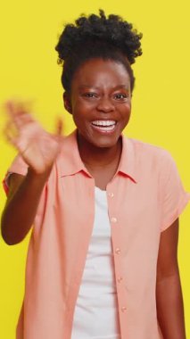 Afro-Amerikan kadın kameraya dostça gülümsüyor, el sallıyor merhaba diyor selam veriyor ya da davetkar bir yüz ifadesiyle veda ediyor. Sarı arka planda izole edilmiş mutlu siyah kız