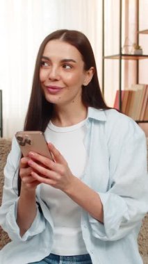 Genç, mutlu bir kadın cep telefonuyla internetten alışveriş yapıyor. Satın alacağı malları seçiyor. Kanepedeki beyaz kız ilgiyle gülümsüyor, egzotik aktivitelerden hoşlanıyor, alışveriş yapıyor.