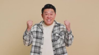 Asyalı orta yaşlı bir adam başarı zaferini kutluyor, doğum gününü kazanıyor, piyango hedefi başarısı, oyun oyunu, iyi haber, zafer. Olgun yetişkin adam bej arka planda kapalı
