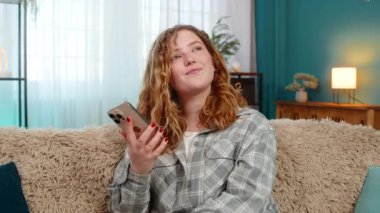 Genç bir kadın akıllı telefondan eski fotoğrafları çekerken mutlu geçmişi hatırlarken kendini nostaljik hissediyor. Kafkasyalı kızıl saçlı kız huzurlu duygusal yüzüyle kanepede oturuyor, sessiz bir anı anı yaşıyor, gülüyor.