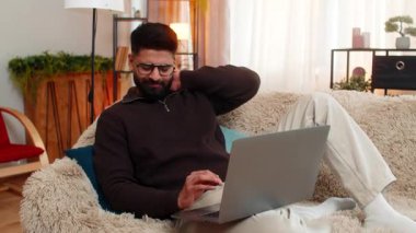 Evde oturan Hintli adam dizüstü bilgisayarın önünde otururken boynu ağrıyor. Uzun dijital çalışmadan sonra yorgun görünüyor. Kanepedeki Arap adam, uzun saatler oturma ve gerilim yüzünden rahatsızlık ve gerginlik gösteriyor.