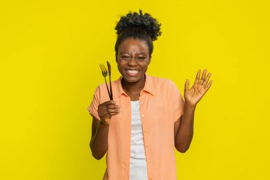 Afro-Amerikalı kadın çatal ve bıçak tutuyor davetkar bir hoş geldin jesti ile, dostluk saçıyor. Sarı arka plandaki siyah kız, ortak yemek deneyiminin zevkini vurguluyor