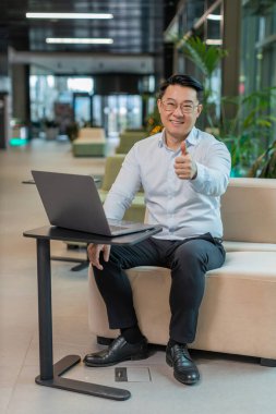 Gibi. Bilgisayarda çalışan mutlu genç işadamı kameraya bakıp olumlu bir işaret veriyor. Modern ofis lobisinde oturan olumlu bir geri bildirim gibi. İş insanları