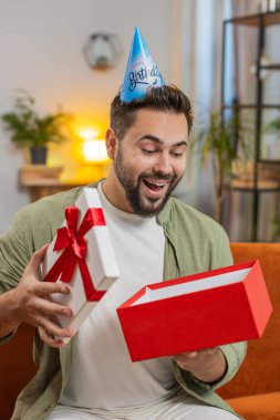 Mutlu genç adam doğum günü şapkası takıyor ve elinde kurdeleyle kutluyor. Beyaz tenli esmer erkek, parti açılışını kutluyor. Kanepede, toplantı odasında hediyeler sunuyor..