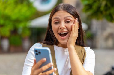 Aman Tanrım. Vay canına. Heyecanlı mutlu, neşeli Avrupalı genç kadın akıllı telefon daktilo kullanıyor kutlama yapıyor açık havada iyi haber kazan. Şehir caddesinde duran yetişkin kız. Kasaba yaşam tarzları