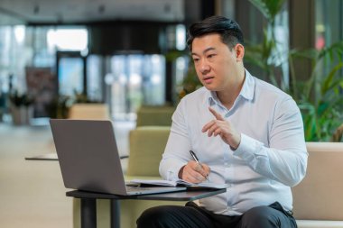Genç Çinli iş adamı, otel lobisindeki internet video dersi veya ofis toplantısına katılırken not alıyor ya da not alıyor. Web kamerasına bakan gömlek destek servislerinde kendine güvenen bir yönetici