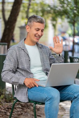 Olgun bir serbest çalışan, şehirde oturuyor, laptop kamerasına bakıyor, arkadaşlarıyla, ailesiyle ya da müşterisiyle görüntülü görüşme yapıyor, uzak sohbetin tadını çıkarıyor. El sallayan adam. Dikey