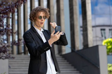 Akıllı telefon kutlaması yapan genç beyaz adam iyi haber kazandı, piyango zaferi kazandı, açık havada dağıttı. Şehir merkezinde kıvırcık saçlı mutlu bir adam. İş adamları yaşam tarzları