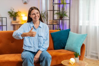 Heyecanlı gülümseyen beyaz kadın portresi onaylayıcı bir şekilde kameraya bakıyor işaret, iyi haber, olumlu geri bildirim gibi. Evdeki oturma odasında kanepede oturan mutlu genç kız..